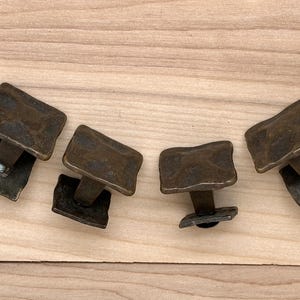 Puede incluir: Cuatro tiradores de armario de color bronce con un dise&ntilde;o rectangular texturizado. Cada tirador tiene una base cuadrada y un tornillo para el montaje. Los tiradores est&aacute;n dispuestos sobre una superficie de madera clara.