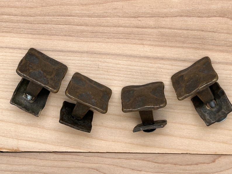 Puede incluir: Cuatro tiradores de armario de color bronce con un dise&ntilde;o rectangular texturizado. Cada tirador tiene una base cuadrada y un tornillo para el montaje. Los tiradores est&aacute;n dispuestos sobre una superficie de madera clara.
