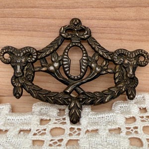 May include: Ornate bronze-toned metal furniture hardware. The piece features a keyhole design, flanked by ram heads and surrounded by a decorative wreath. The hardware is likely for a drawer or cabinet.