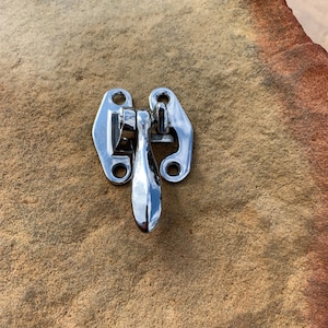 May include: A silver-toned metal latch with a lever handle. The latch is mounted on a brown textured surface.