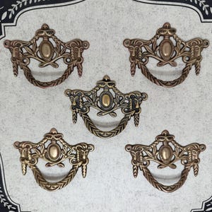 May include: Five antique bronze drawer pulls with ornate floral and face designs. Each pull has a curved handle and an oval center. The pulls are arranged on a light gray background with a decorative border.