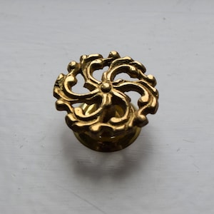 May include: A gold-toned metal cabinet knob with a decorative, intricate, floral design.
