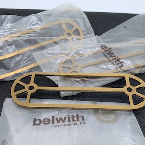 May include: Four gold-colored metal door pulls with a decorative design. The pulls are packaged in clear plastic bags with the brand name "belwith international, ltd." printed on the bags.