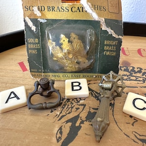 May include: Three antique brass cabinet door pulls, two with a ring handle and one with a decorative scroll design. The pulls are arranged on a wooden surface with letter tiles spelling out the words "A", "B", and "C".