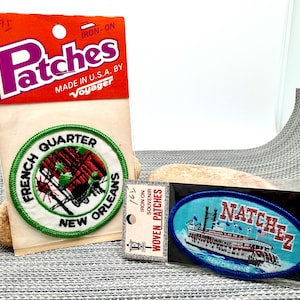 May include: Two iron-on embroidered patches in original packaging. One circular patch features a red building with green trees and the text French Quarter New Orleans. The other oval patch depicts a steamboat with the text Natchez New Orleans.
