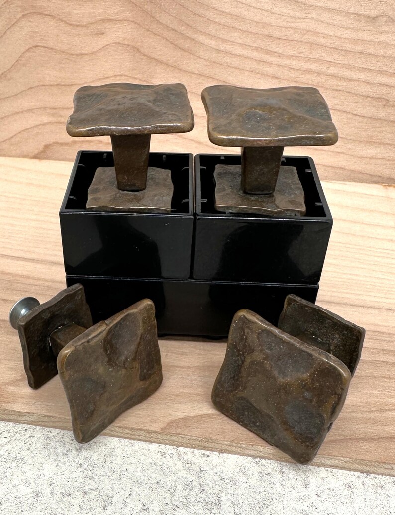 Puede incluir: Juego de tiradores cuadrados de armario en tono bronce con superficie texturizada. Dos tiradores se muestran sobre cajas negras, con tiradores adicionales cerca. Los tiradores tienen un aspecto r&uacute;stico y envejecido.