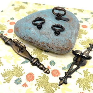 May include: Assortment of antique-style cabinet hardware in a brass finish. The hardware includes a long, ornate pull, a small, ornate knob, and two small, ornate pulls with a keyhole design.