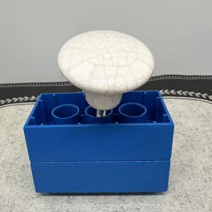May include: A white ceramic knob with a crackle glaze finish is attached to a bright blue rectangular base. The knob is round and has a slightly domed shape. The base has three circular openings. The background is a light gray.