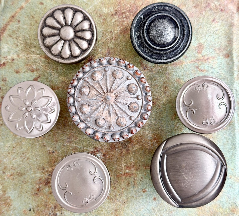 Może przedstawiać: Asortyment siedmiu dekoracyjnych gałek do szafek w r&oacute;żnych wzorach i wykończeniach. Gałki obejmują wzory kwiatowe, geometryczne i ozdobne, w kolorach od srebrnego do ciemnoszarego. Gałki te są przeznaczone do mebli lub wystroju domu.