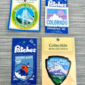 May include: Four embroidered iron-on patches featuring Colorado landmarks. One round Royal Gorge patch, one square Colorado patch with a purple sun, one Winter Park patch with a skier, and one shield-shaped Pikes Peak patch with 14,110 FT text.