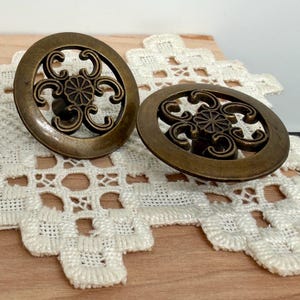 May include: Two antique bronze-colored cabinet knobs. Each knob features an oval shape with an openwork design. The design includes a central floral element surrounded by swirling, curved lines. The knobs are set against a white lace doily and a wooden surface.