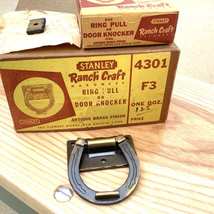 May include: Antique brass door knocker or ring pull in the shape of a horseshoe. The knocker is on a wooden surface with a box labeled "Stanley Ranch Craft Hardware 4301 F3 One Doz. 1.25 Price".