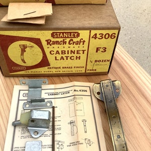 May include: A box of Stanley Ranch Craft cabinet latches in antique brass finish. The box is yellow with red and black text. The box is labeled 4306 F3 and says "One Dozen". The box is sitting on a wooden surface. There is a close-up of the latch and instructions for installation.