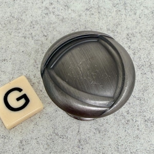 Może przedstawiać: Szary, metalowy uchwyt do szafki o okrągłym kształcie i szczotkowanym wykończeniu. Uchwyt ma wypukły, zakrzywiony wz&oacute;r. Obok uchwytu znajduje się płytka do Scrabble z literą "G".