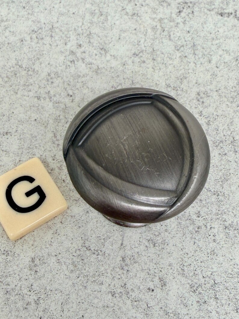 Może przedstawiać: Szary, metalowy uchwyt do szafki o okrągłym kształcie i szczotkowanym wykończeniu. Uchwyt ma wypukły, zakrzywiony wz&oacute;r. Obok uchwytu znajduje się płytka do Scrabble z literą "G".