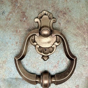 May include: An antique-style door knocker in a bronze finish. The knocker features a decorative backplate with an ornate design and a curved handle. The overall design is symmetrical and classic, suitable for a front door.