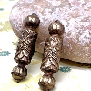 May include: A pair of antique gold-toned metal earrings with intricate floral designs. The earrings are cylindrical in shape with a rounded top and bottom.