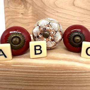 May include: Three decorative cabinet knobs. Two are a glossy red with brass hardware, and one is white with a floral design. The knobs are positioned with lettered tiles spelling out "A", "B", and "C".