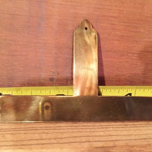 May include: A brass T-shaped plaque with rounded edges and a hole at the top. The plaque is approximately 13 cm long and 4 cm wide.