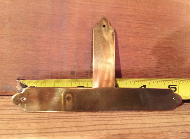 May include: A brass T-shaped plaque with rounded edges and a hole at the top. The plaque is approximately 13 cm long and 4 cm wide.