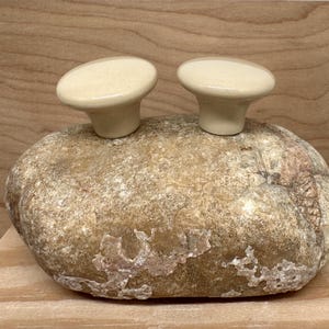 May include: Two off-white ceramic cabinet knobs with a rounded oval shape are displayed on a large, textured, light brown rock. The knobs have a smooth, glossy finish and a short, cylindrical base.