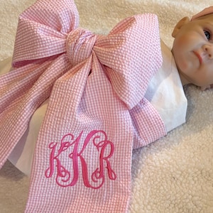 May include: A large pink and white gingham seersucker bow featuring a decorative hot pink embroidered monogram with the letters KKR. The bow is attached to a white fabric base and is positioned next to a doll wearing a matching pink headband.