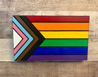 May include: A wooden sign featuring a rainbow flag design. The flag has eight stripes in the colours of red, orange, yellow, green, blue, purple, brown and pink. The stripes are arranged in a diagonal pattern, with the brown and pink stripes forming a chevron shape in the top left corner.