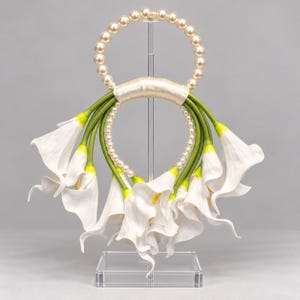 May include: A circular handbag handle featuring a ring of cream-colored faux pearls at the top and a smaller ring of pearls at the bottom, connected by green stems of white calla lily flowers wrapped in a smooth, light beige satin ribbon.