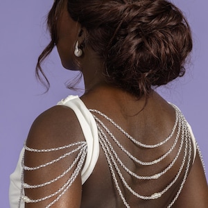 May include: A woman's back is shown, wearing a white dress and a shoulder necklace. The necklace is made of multiple strands of clear beads and pearls. The woman has dark hair styled in an updo, and is wearing pearl earrings and a headpiece.