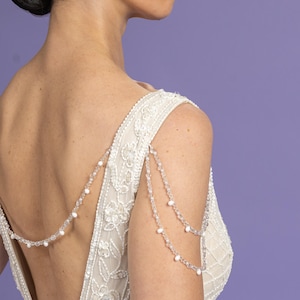 May include: A beaded shoulder necklace with a low-back wedding dress. The dress is ivory with floral embroidery and sequin details. The shoulder necklace features clear beads and small white pearls, draping elegantly across the shoulders and back.