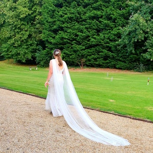 Puede incluir: Una novia con un vestido de novia blanco y un velo largo y fluido camina por un camino de grava. El camino está rodeado de hierba verde y árboles.