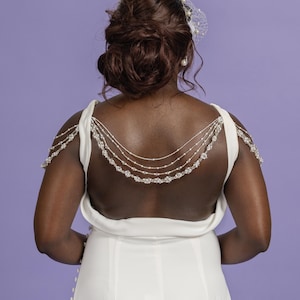 May include: A white backless wedding dress with a beaded shoulder and back detail. The dress has a simple, elegant design. The model's hair is styled in an updo, and a veil is attached to the hair.