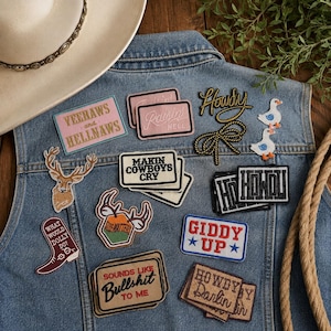 May include: A collection of embroidered patches on a blue denim vest. Patches include text like YEEHAWS AND HELLNAWS, RAISIN HELL, MAKIN COWBOYS CRY, HOWDY, GIDDY UP, WHAT WOULD DOLLY DO, SOUNDS LIKE BULLSHIT TO ME, and HOWDY DARLIN, plus deer and duck motifs.