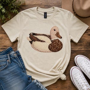 Könnte beinhalten: Beiges T-Shirt mit einem Patchwork-Enten-Design in Braun- und Cremetönen. Die Ente hat einen karierten Kopf, einen floral gemusterten Körper und einen gepunkteten Flügel. Das Shirt wird mit blauen Jeans und weißen Turnschuhen kombiniert, was einen lässigen Look ergibt.