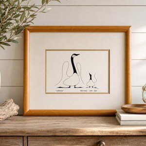 May include: Framed art print featuring a minimalist illustration of two geese in black and white. The artwork is displayed in a wooden frame with a cream-colored mat. The print is set on a wooden surface with decorative elements.
