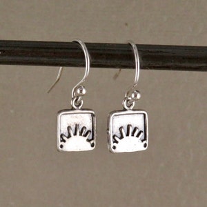 May include: A pair of silver earrings with a square design featuring a stylized sun graphic.