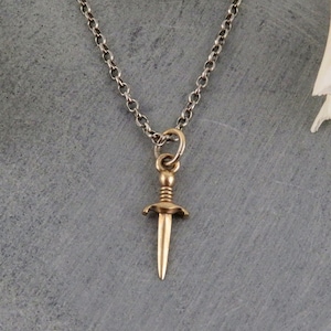 May include: A silver chain necklace with a gold dagger pendant. The dagger is detailed with a small ball at the top and a flared base.