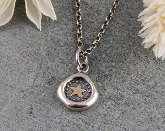Könnte beinhalten: Eine silberne Halskette mit einem kleinen, unregelmäßigen Anhänger mit einem goldenen Stern. Der Anhänger ist von kleinen Punkten umgeben. Die Halskette wird auf einer grauen Oberfläche mit weißen Trockenblumen präsentiert.
