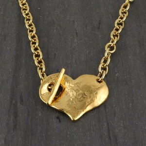 May include: A gold-colored necklace featuring a heart-shaped pendant. The pendant has a textured surface and is connected to a chain. The necklace has a toggle clasp. The chain is approximately 18 inches long.