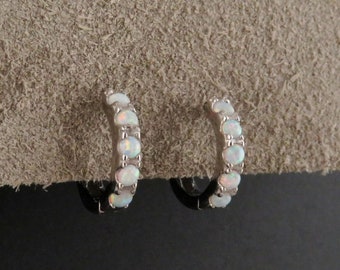 May include: A pair of small hoop earrings. Each earring is silver-toned and features a row of oval-shaped, white opal stones. The earrings are set against a textured, light brown surface and a black background.
