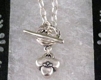 May include: A silver-toned necklace with a toggle clasp and a clover-shaped pendant. The chain is made of small, linked circles. The pendant has a detailed, three-dimensional design. The necklace is displayed against a textured, light-colored background.