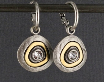 Op de afbeelding: Een paar zilveren oorbellen met gouden accenten en een helder kristal in het midden van elke oorbel. De oorbellen hebben de vorm van een cirkel met een getextureerd oppervlak.