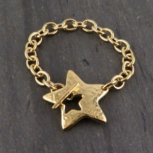 May include: Gold bracelet with a star-shaped charm. The charm has a hammered texture and a cutout design. The bracelet has a chain link design and a toggle clasp. The star charm is approximately 3.8 cm wide.