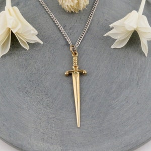 May include: A silver chain necklace with a gold sword pendant. The pendant is a simple, stylized design with a pointed tip. The necklace is displayed on a gray surface with white flowers.