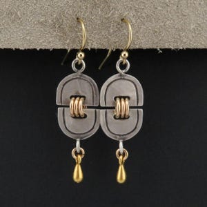Puede incluir: Un par de pendientes de plata y oro con un diseño geométrico. Los pendientes tienen forma de lágrima con una pequeña perla dorada en la parte inferior.