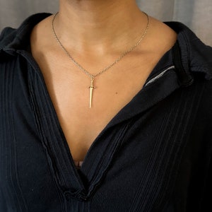 Peut inclure: Un collier avec un pendentif en forme de dague dor&eacute;e sur une cha&icirc;ne en argent. Le collier est port&eacute; par une personne avec un chemisier noir.