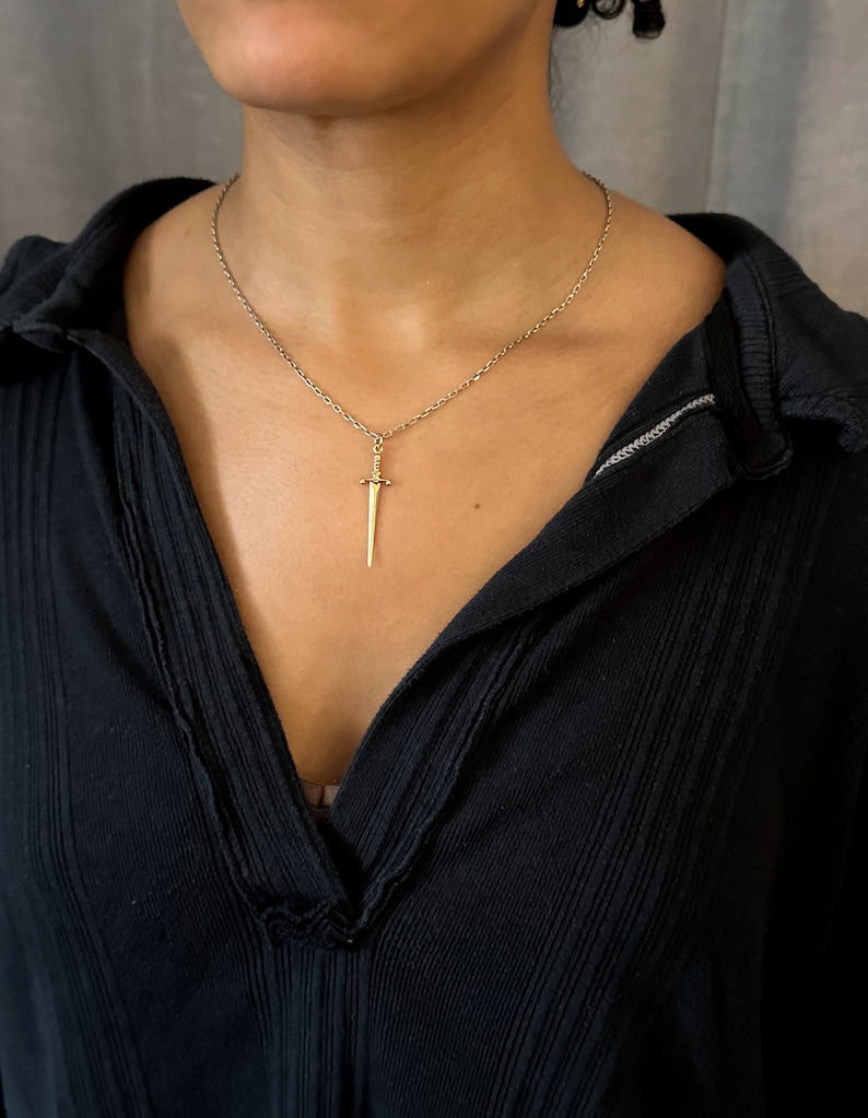 Peut inclure: Un collier avec un pendentif en forme de dague dor&eacute;e sur une cha&icirc;ne en argent. Le collier est port&eacute; par une personne avec un chemisier noir.
