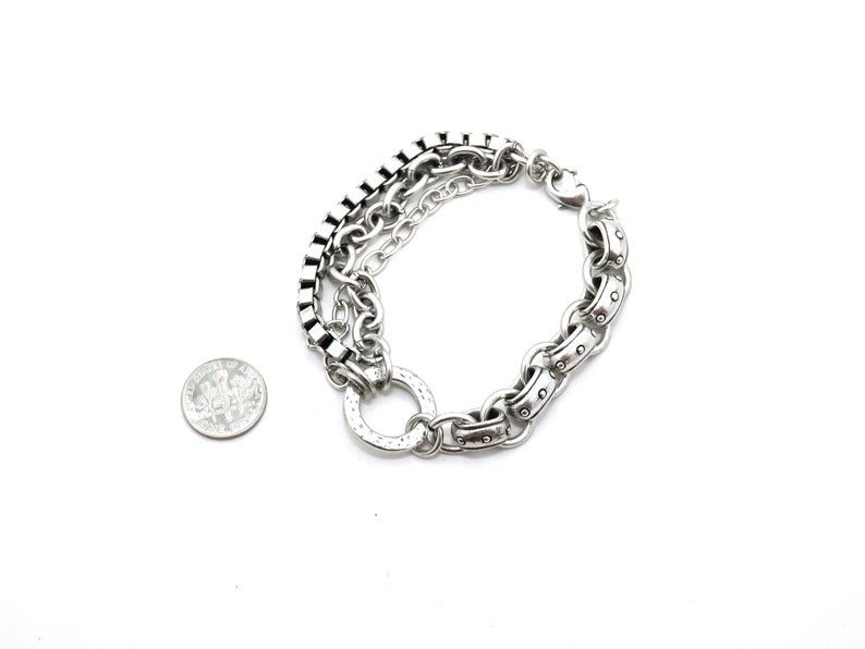 May include: Silver-toned bracelet featuring a multi-chain design with a variety of link styles. The bracelet includes a square link chain, a classic chain, and a larger, studded chain. A dime is included for size reference.