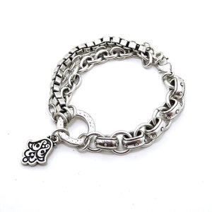 May include: Silver-toned bracelet featuring a layered design with a mix of chain styles. One layer has square links, while another has rounded links with decorative accents. A charm in the shape of a hand with ornate details hangs from the bracelet.