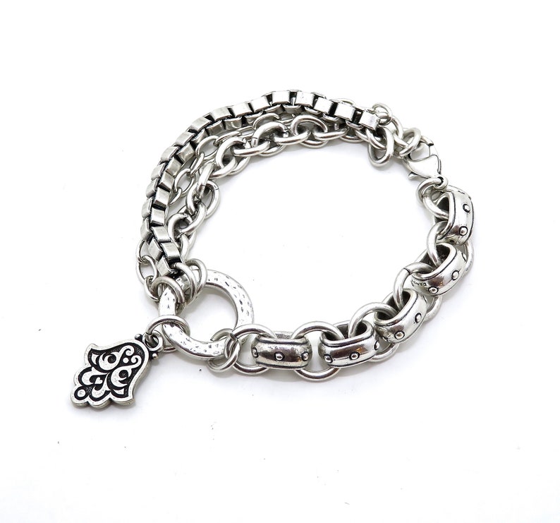 May include: Silver-toned bracelet featuring a layered design with a mix of chain styles. One layer has square links, while another has rounded links with decorative accents. A charm in the shape of a hand with ornate details hangs from the bracelet.